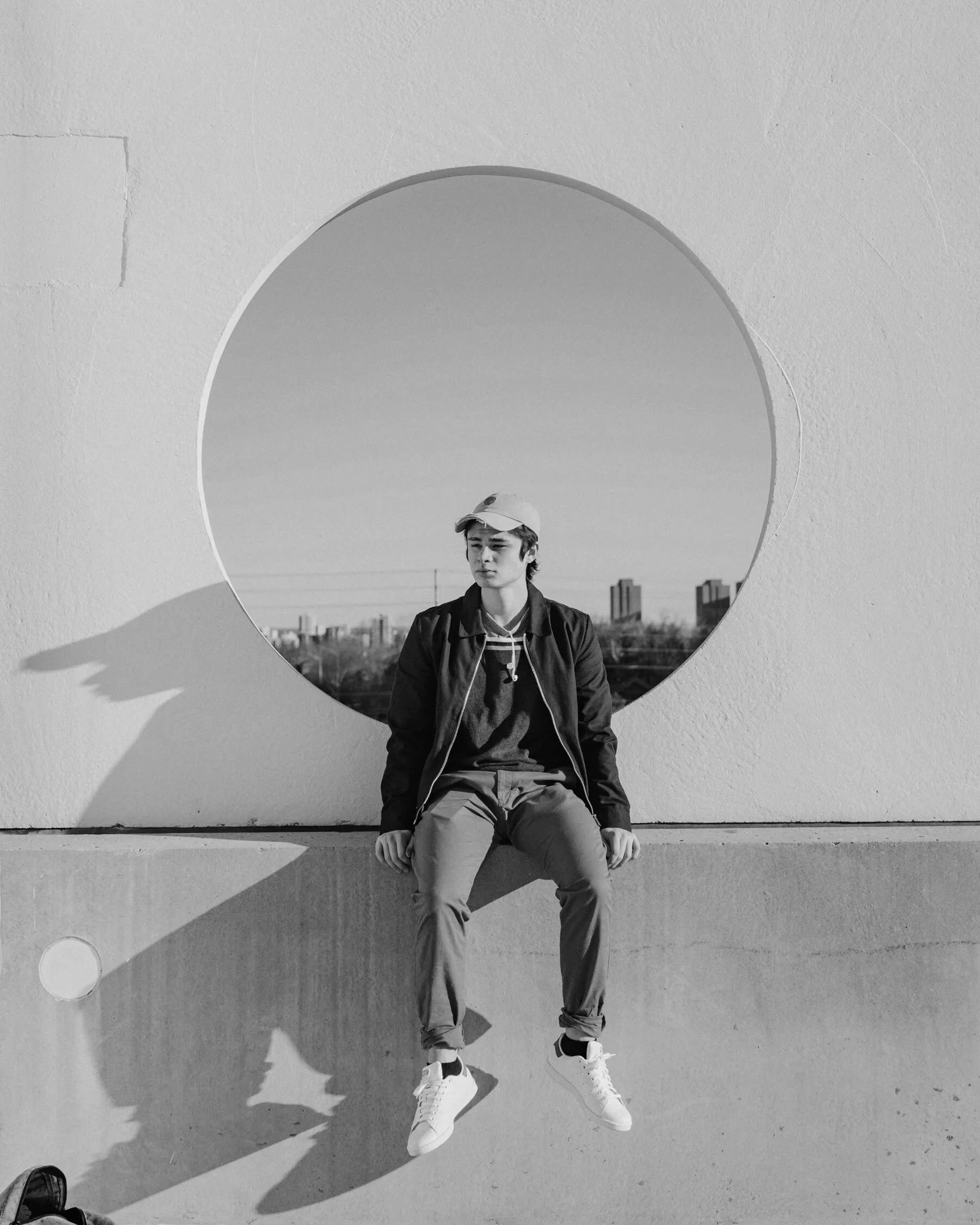 A young man sits on a ledge in front of a circular window, his shadow cast long across the wall