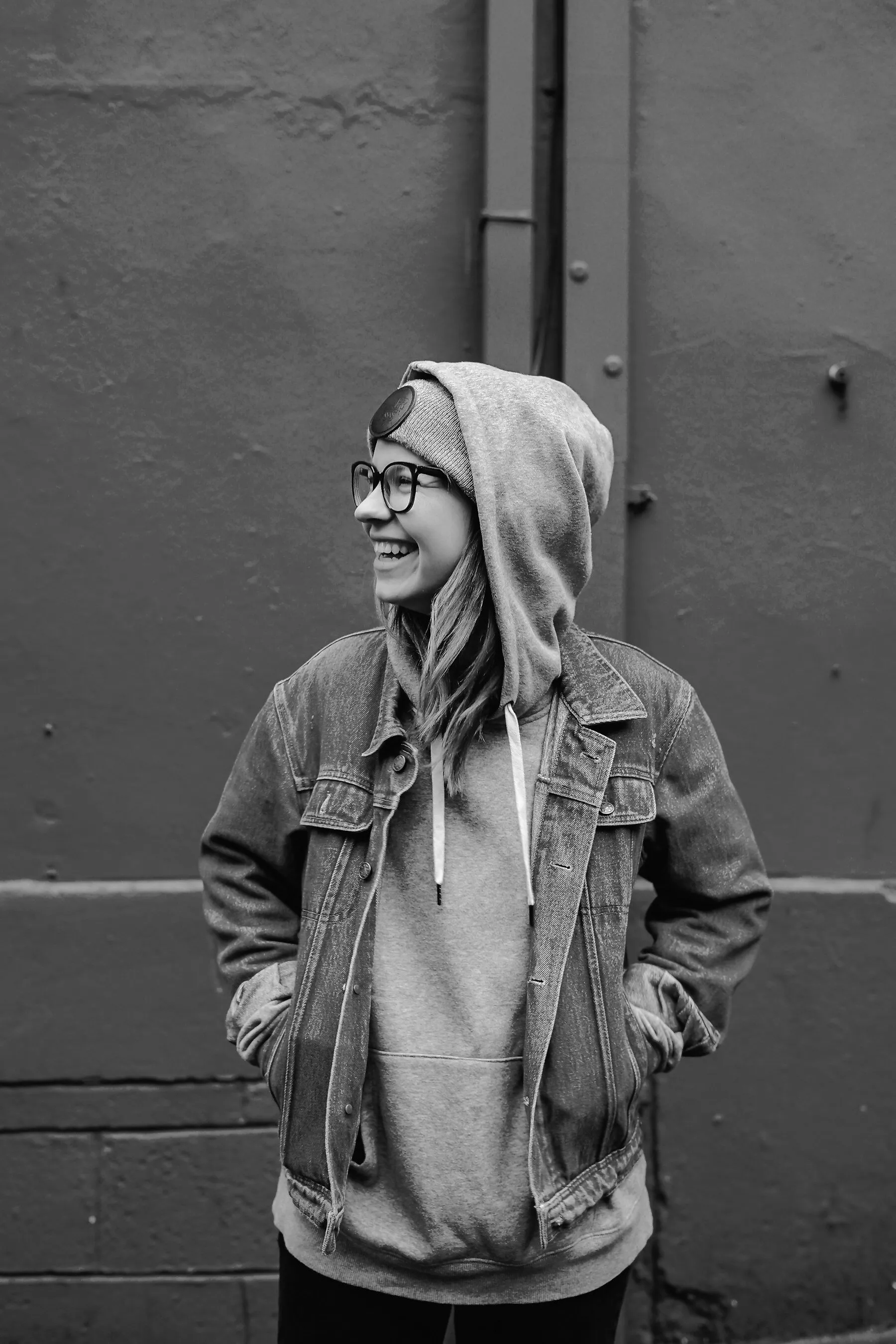A man in a hoodie and denim jacket laughing against a wall